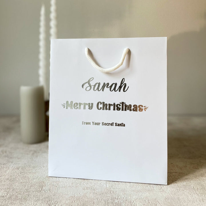 A white gift bag with rope handles, featuring Secret Santa Gift Bags personalized text 'Sarah' and 'Merry Christmas' from 'Your Secret Santa'.