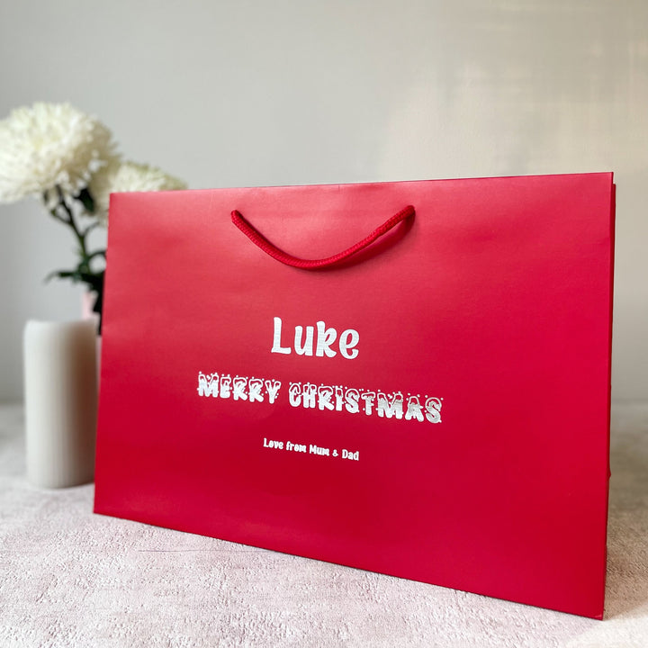 A personalised Christmas gift bag for children with the name 'Luke' printed on it, along with the message 'Merry Christmas' in festive lettering, placed on a table with a vase and flowers in the background.