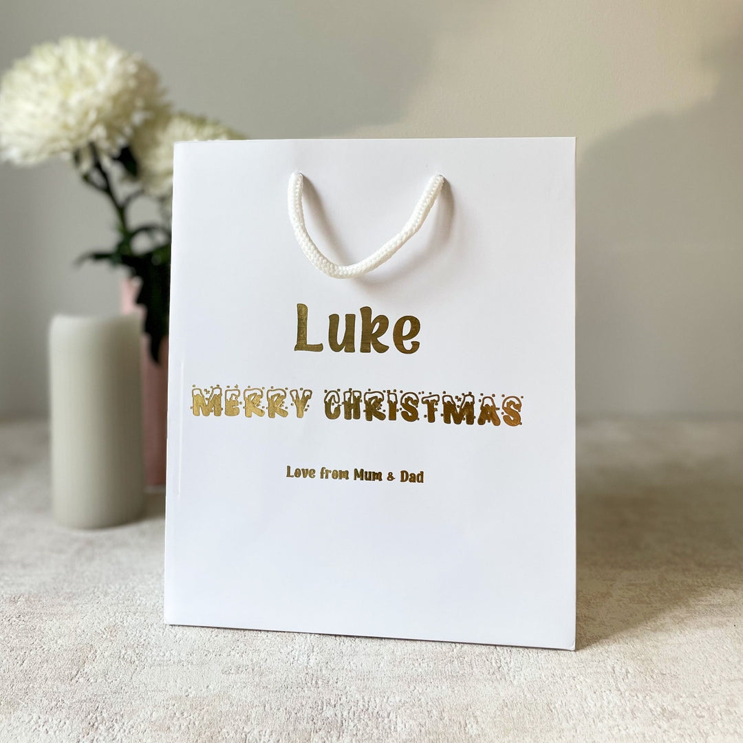 A personalised Christmas gift bag for children with the name 'Luke' printed on it, along with the message 'Merry Christmas' in festive lettering, placed on a table with a vase and flowers in the background.