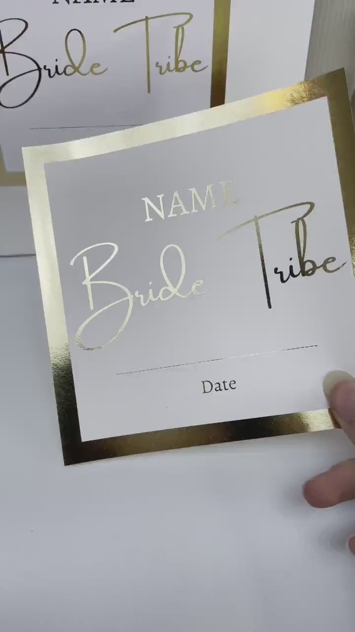 A white personalized gift bag for a Hen Party, featuring a gold-framed label with customizable fields for "NAME" and "Date." The text "Bride Tribe" is prominently displayed in a stylish font. The bag is surrounded by a rose and elegant decorative items.