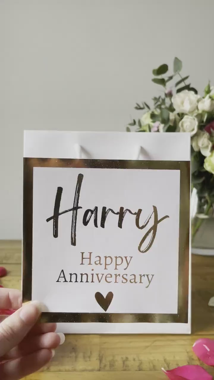 A white Anniversary gift bag with a 'Happy Anniversary' label featuring the name 'Harry' in elegant, gold script. The label is bordered with a gold square frame and a small heart at the bottom. The bag is placed on a wooden table, with a pink rose in the foreground and a soft floral arrangement visible in the background.