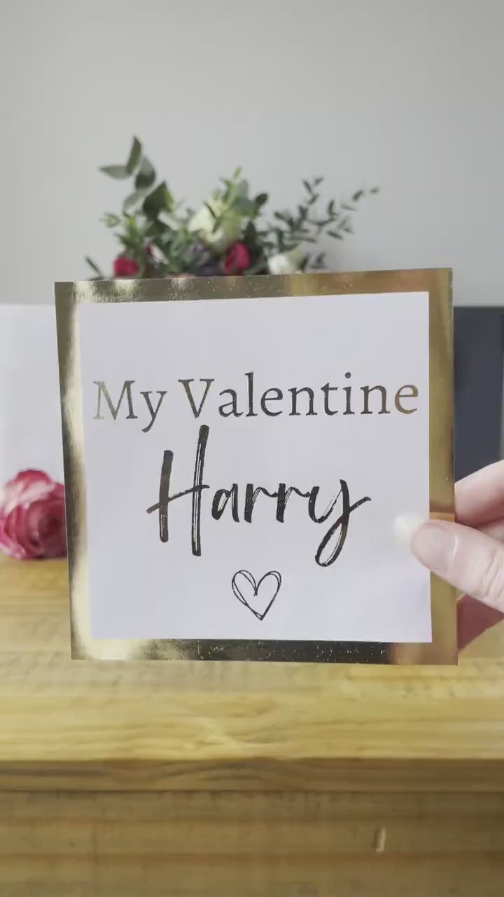 "Valentine's gift bag with the text 'My Valentine Harry' on a white bag, surrounded by rose petals, a glass of champagne, and a vase of flowers in the background."