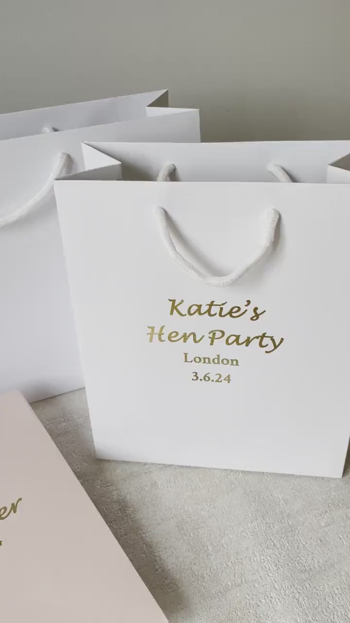 A white hen party gift bag with gold text and detailing, displayed next to a glass of champagne and decorative foliage. 